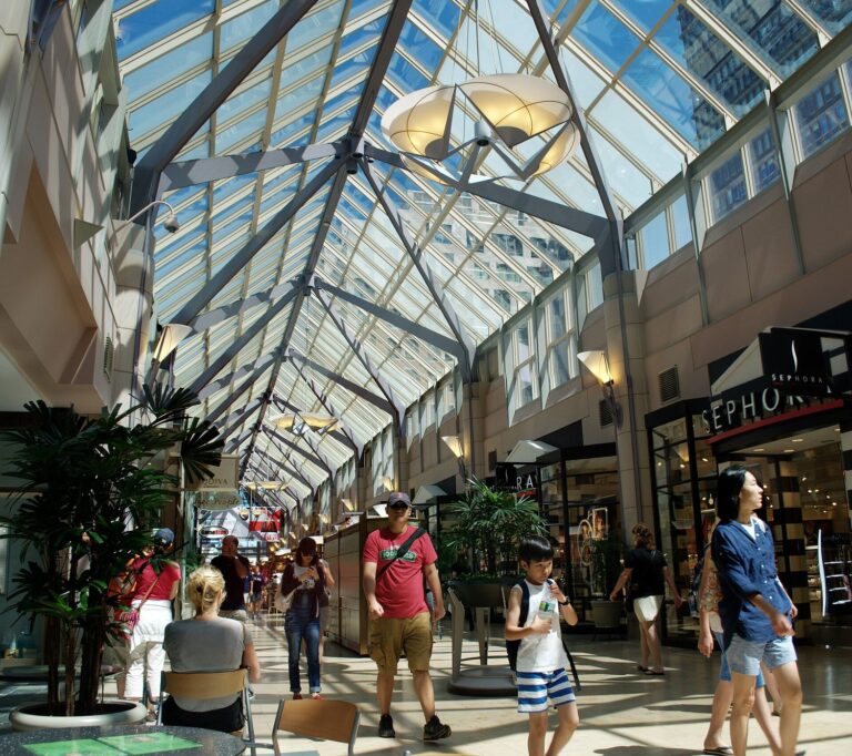 The glass-roofed arcade of the Shops at Prudential Center in Boston, with shoppers walking beneath a steel-and-glass ceiling past store fronts and potted palms.