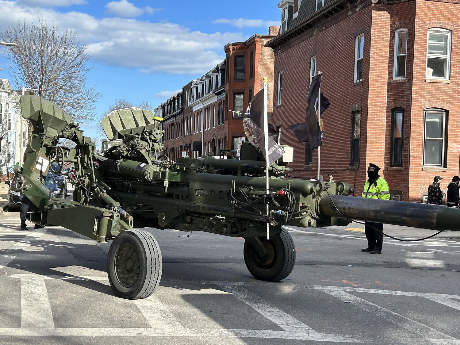 A towed military howitzer being pulled along a South Boston street during the St Patrick's Day parade, with brick row houses and a police officer at the crossing.
