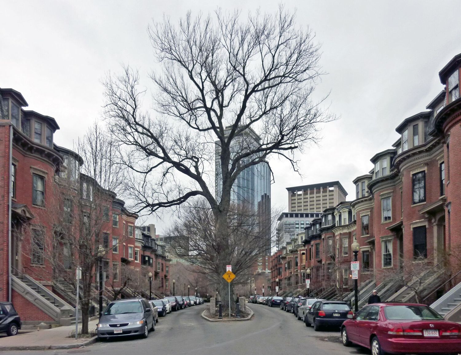 A South End residential street lined with bowfront brick row houses and stone stoops, a tree-planted median down the middle and the Prudential Tower visible beyond.