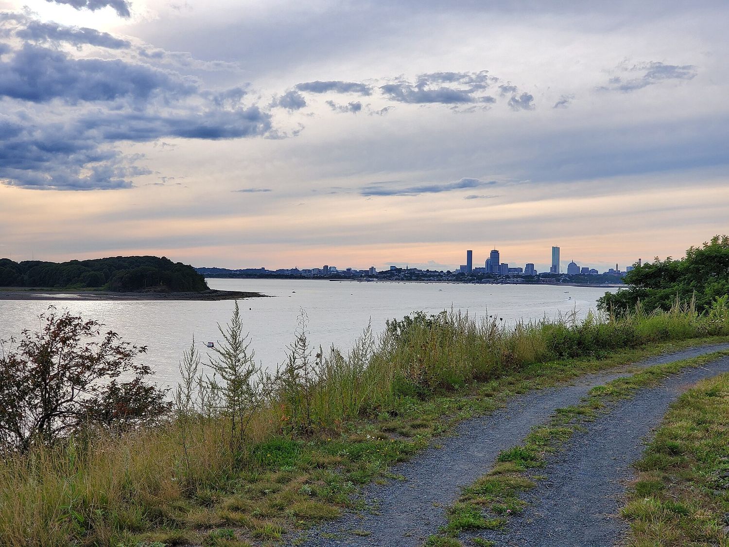A gravel path running along the grassy shoreline of Spectacle Island in Boston Harbor at dusk, with the Boston skyline low on the horizon across the water.