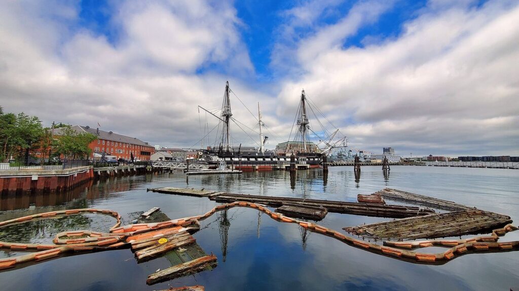 USS Constitution moored at the Charlestown Navy Yard, her masts reflected in still harbor water.