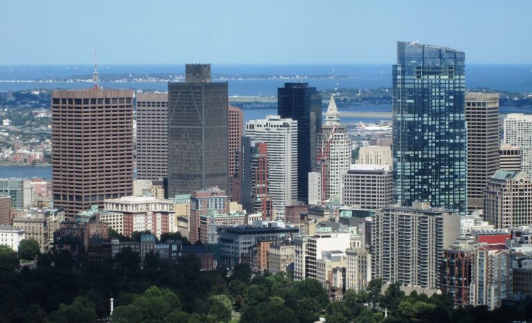 Downtown Boston's towers seen from the top of the Prudential Tower, with the Custom House clock tower among them, Logan Airport, the harbor and the Harbor Islands behind, and the trees of the Common in the foreground.